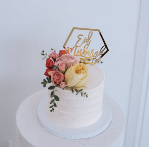 Round white cake with roses and greenery. Gold hexagon topper reading "Eid Mubarak" is the star product of this image.