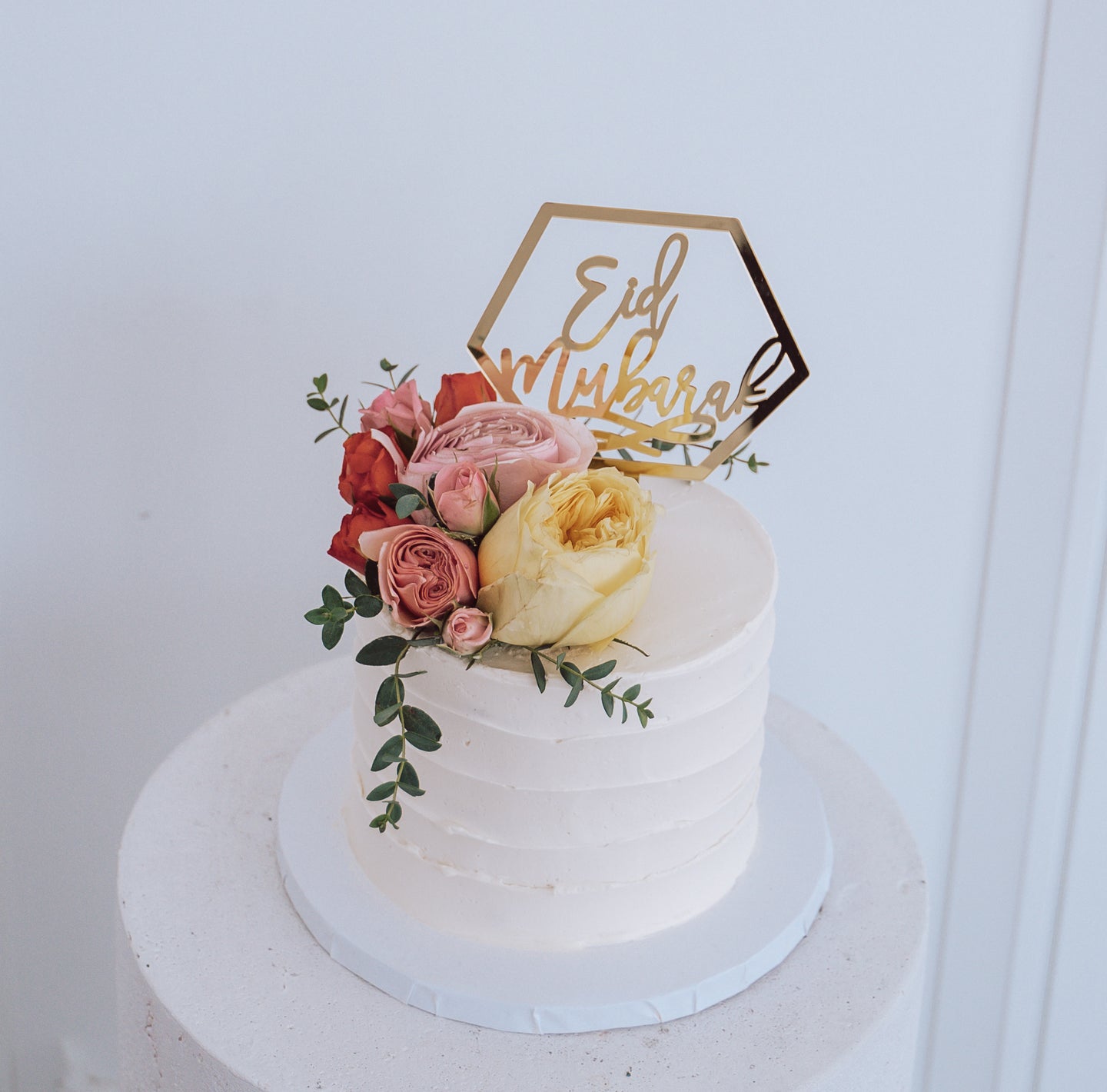 Round white cake with roses and greenery. Gold hexagon topper reading 